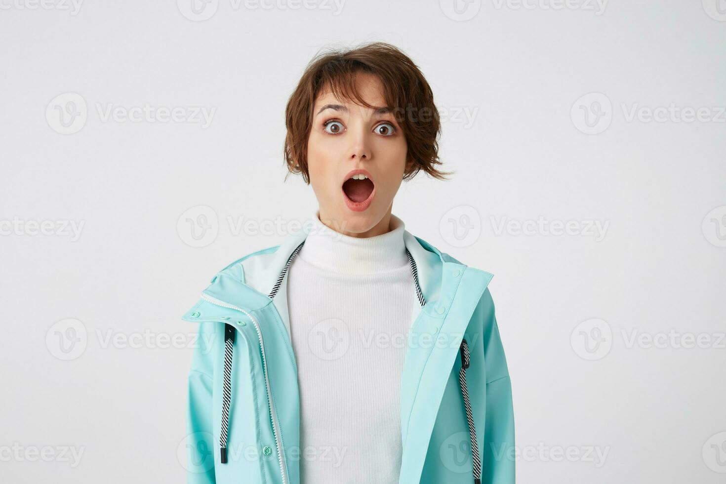 Close up of wondered short-haired young woman in white golf and blue rain coat, stands over white background with wide open mouth and surprised expression, looks at the camera with wide open eyes. photo