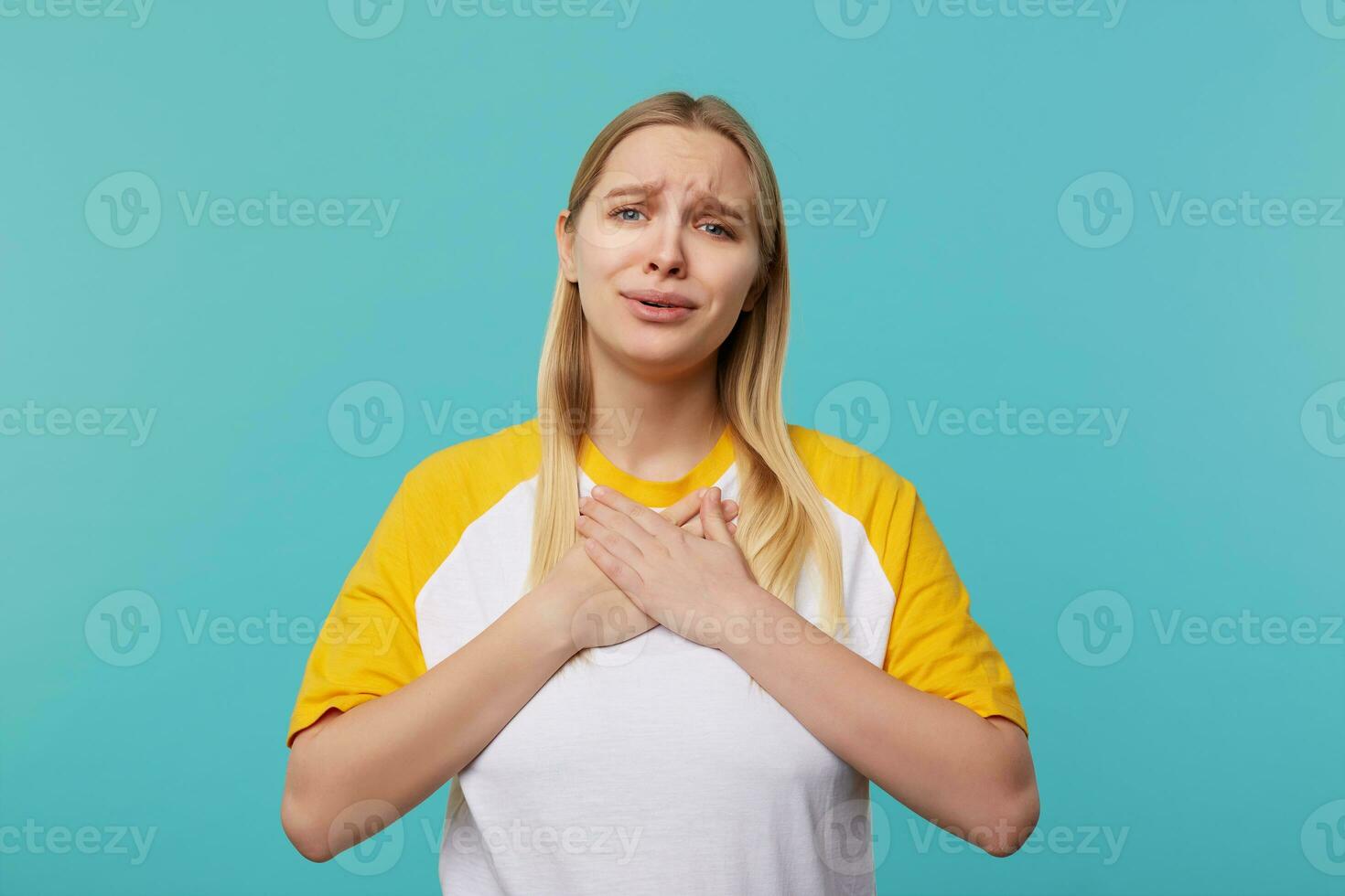 Sweet young pretty blue-eyed long haired blonde woman holding palms on her chest while looking emotionally at camera, wearing casual clothes while posing over blue background photo