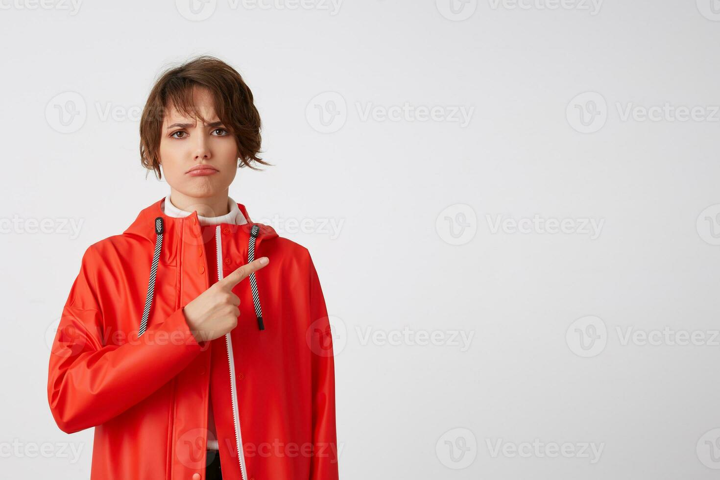 Young unhappy cute short haired lady dressed in red rain coat, looking with sad expression, wants to draw your attention to the copy space, points fingers to the right. Standing over white background. photo