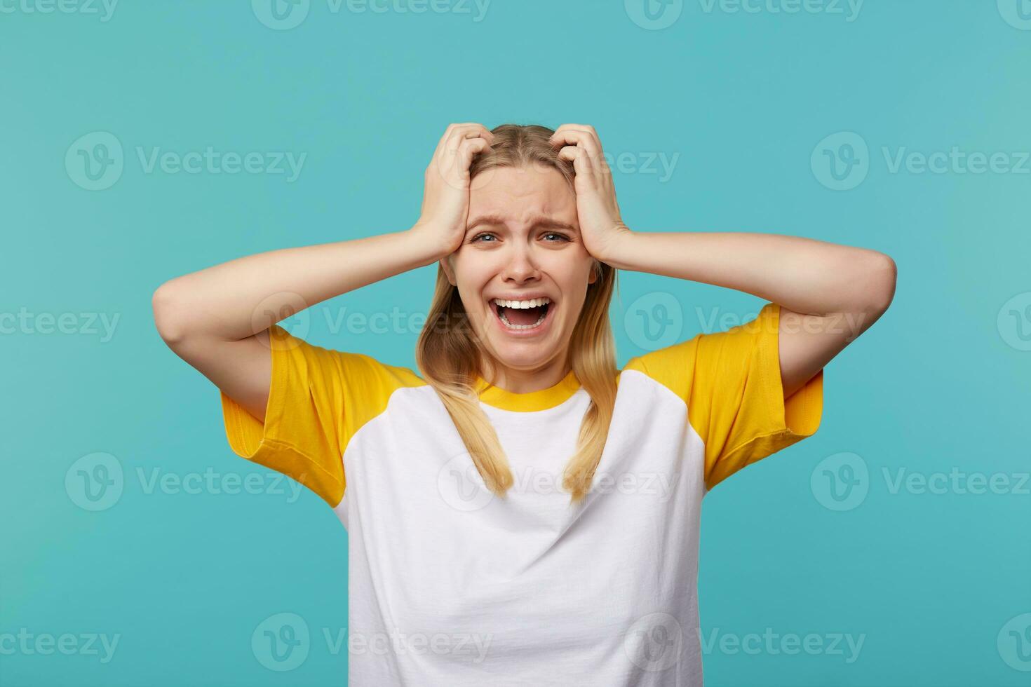 Unhappy young blue-eyed blonde lady with long hair keeping raised hands on her head and looking desperately at camera, standing against blue background photo