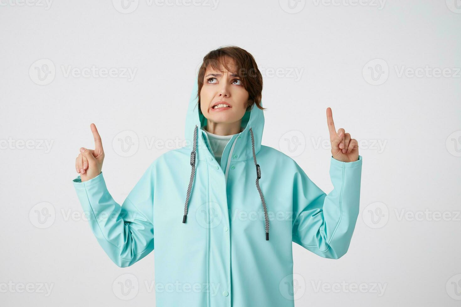 Cute disgusting short-haired curly woman in blue rain coat, hiding under the hood from rain and looking up, wants to draw you attention to the copy space under shes head, stands over white background. photo
