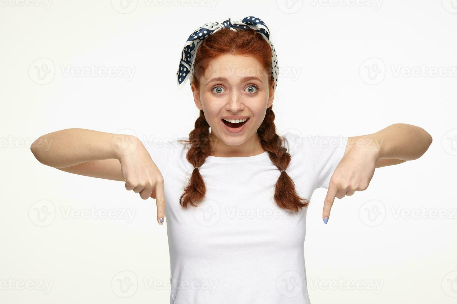 Indoor portrait of young ginger female posing over white background points with a finger to bottom side of the picture photo