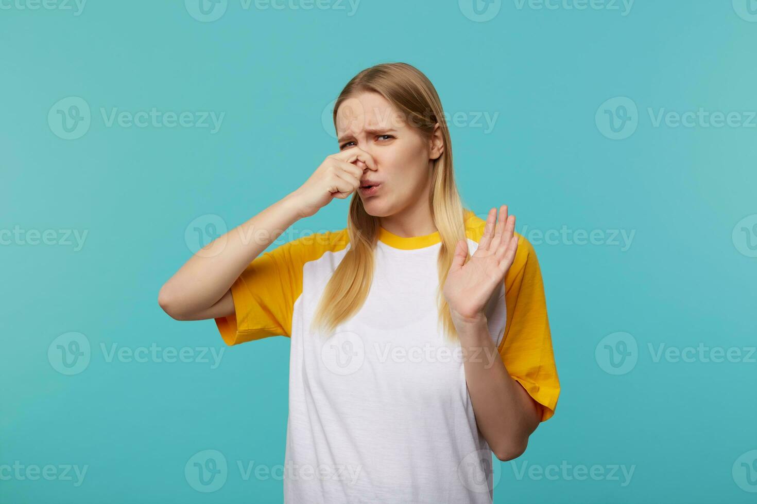 Displeased young attractive long haired blonde female closing her nose with raised hand and frowning face while feeling bad smell, posing over blue background photo