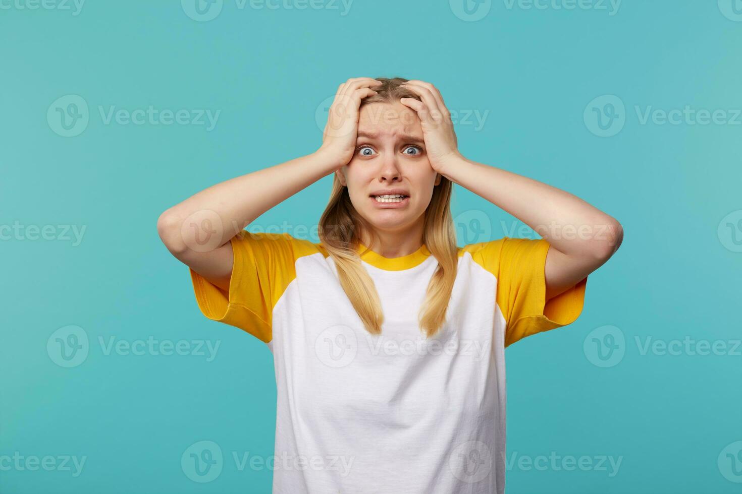 Depressed young blue-eyed fair-haired woman with casual hairstyle clutching her head with raised hands and looking scaredly at camera, isolated over blue background photo
