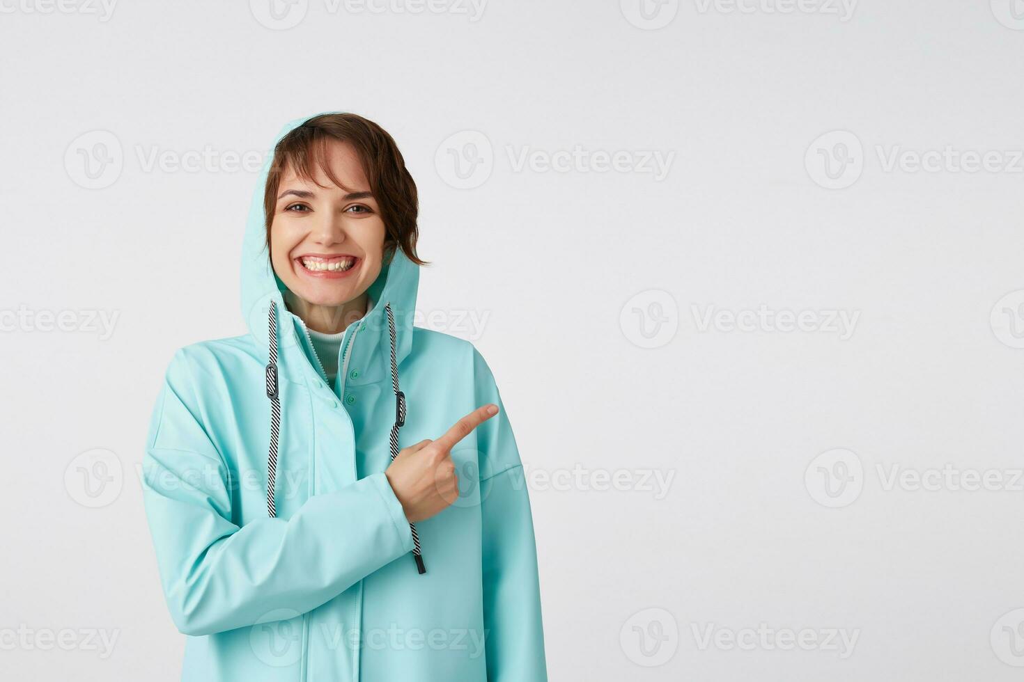Cute smiling short-haired curly lady in blue rain coat, wants to draw you attention to the copy space on the right side and pointing at him, stands over white background. photo