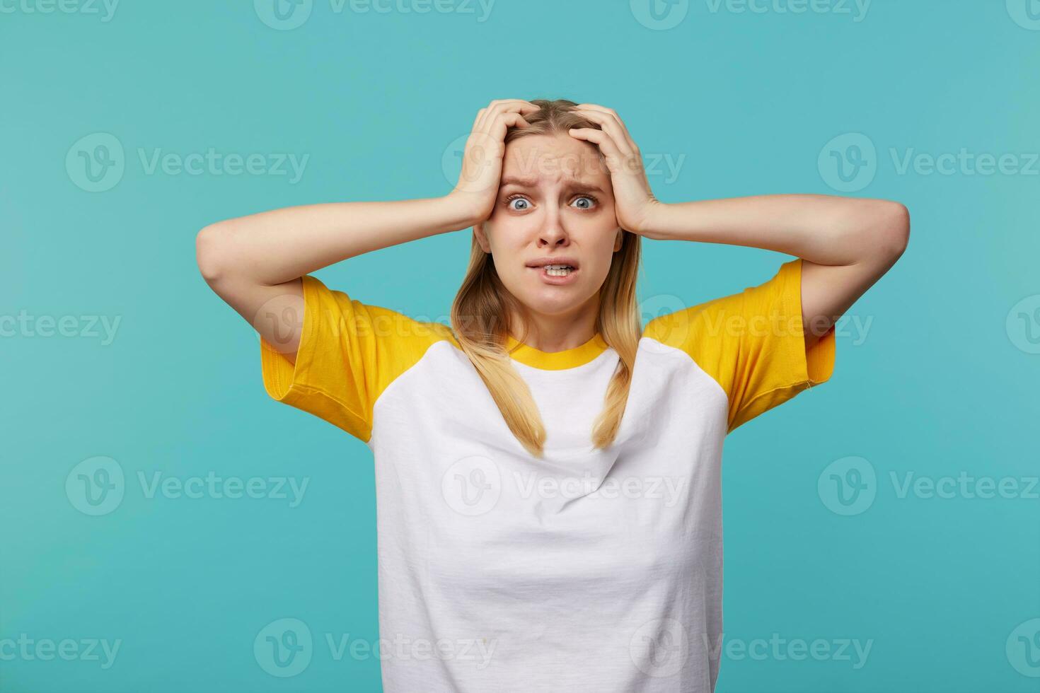Afraid young attractive long haired blonde woman with natural makeup holding her head with raised hands and biting scaredly underlip, posing over blue background photo