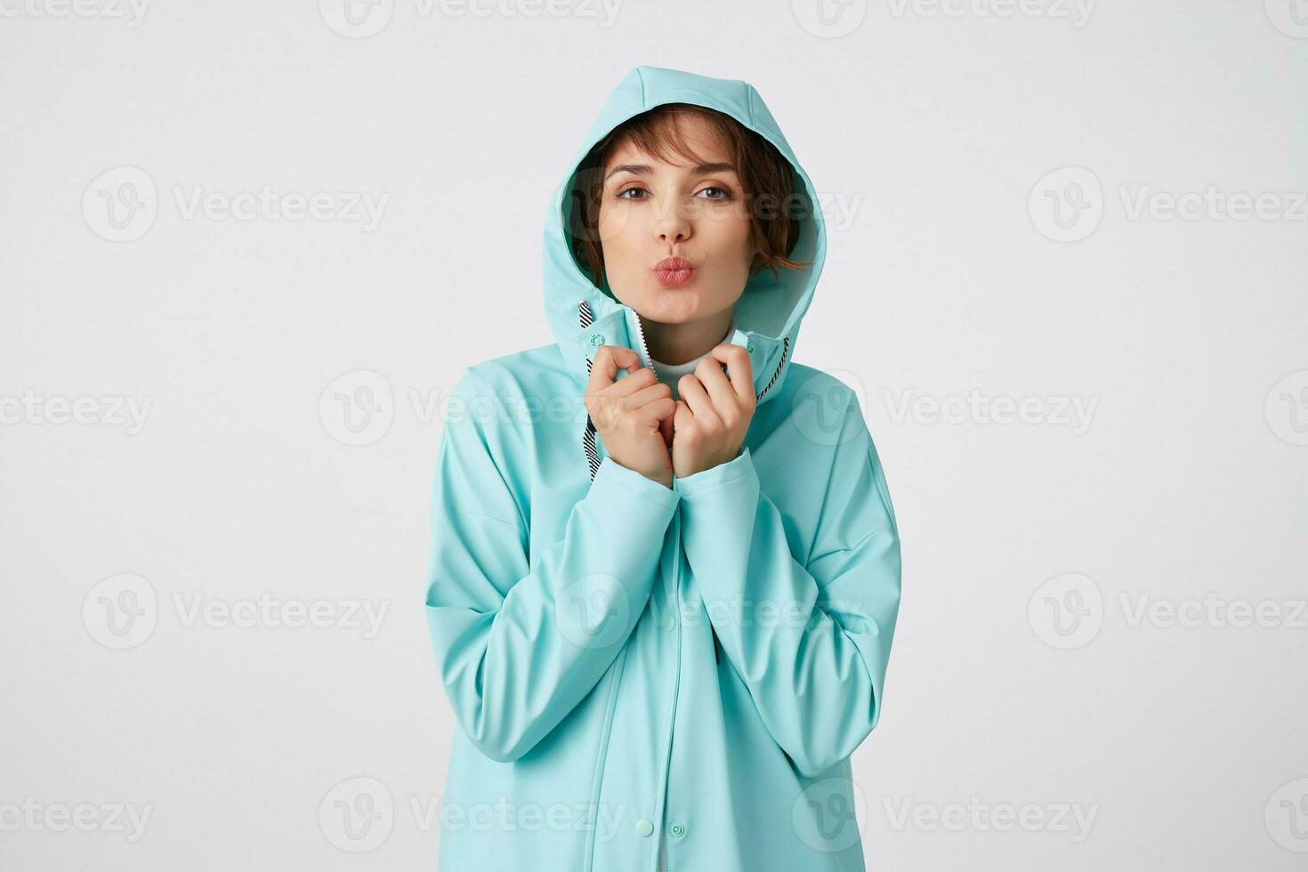 Portrait of positive young nice lady in blue rain coat, with a hood on his head, looks at the camera with happy expressions, send kiss at the camera, stands over white wall. photo