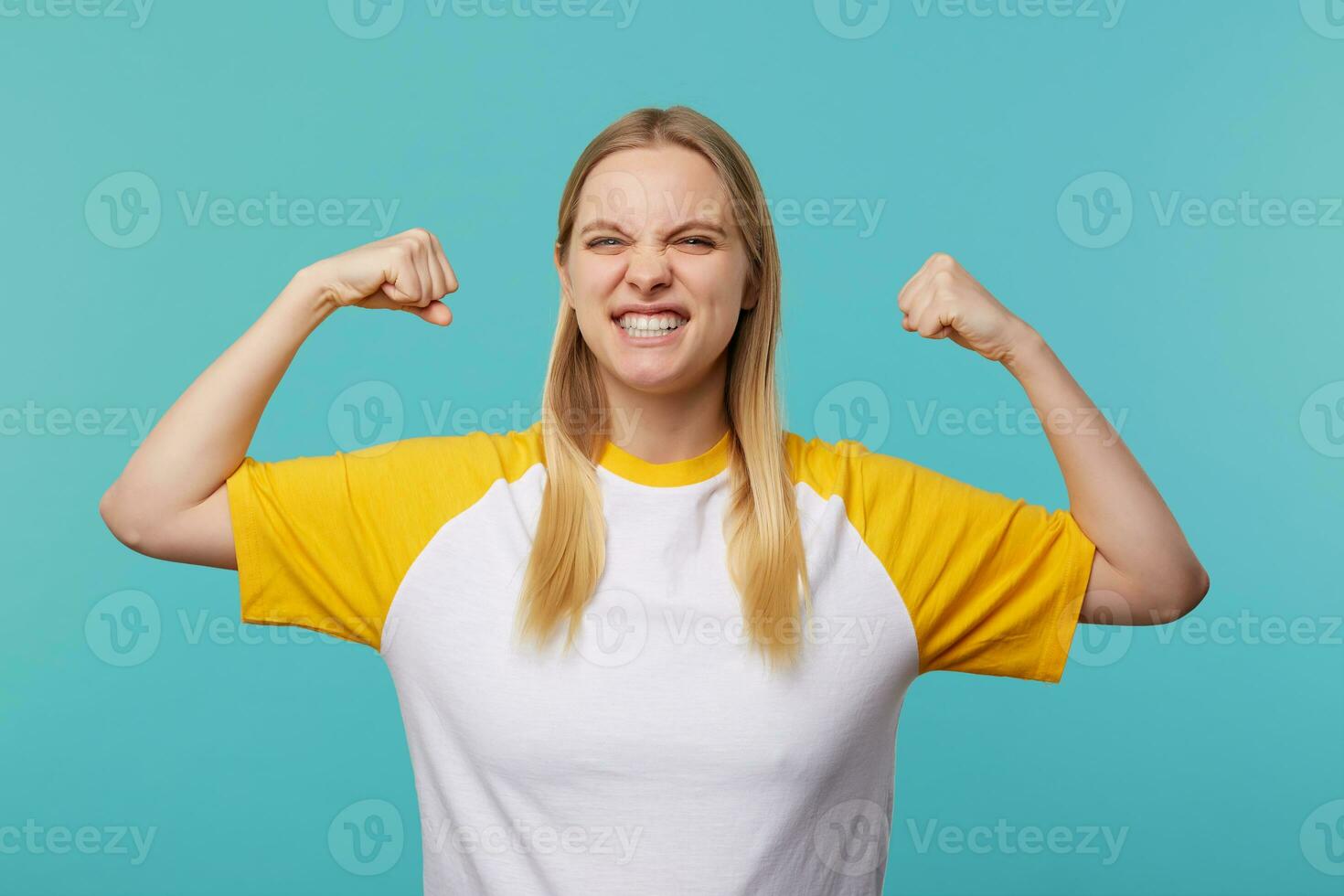 Portrait of strong young lovely long haired blonde woman raising her hands while demonstrating power and frowning her face, isolated over blue background photo
