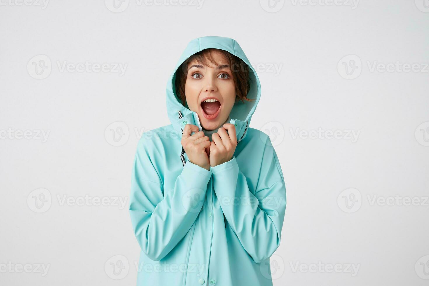 Photo of happy amazed young beautiful woman in blue rain coat, with a hood on his head, looks at the camera with surprise expressions, stands over white wall.