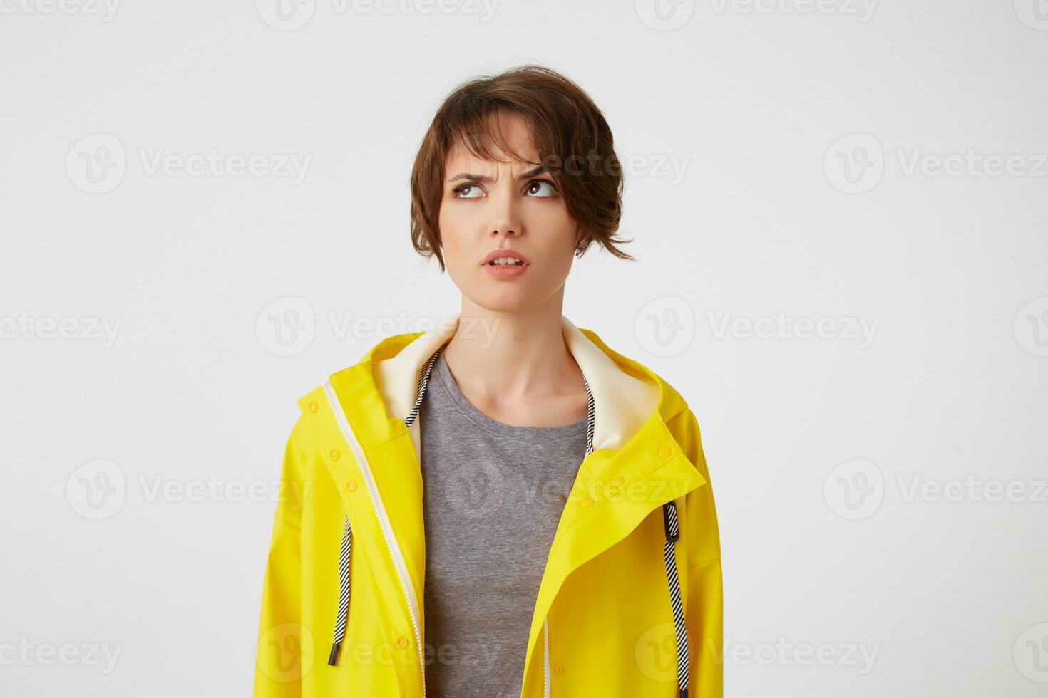 Photo of young thinking short-haired lady in yellow rain coat, looks discontent and doubting, frowning looks up at the left side, stands over white background.