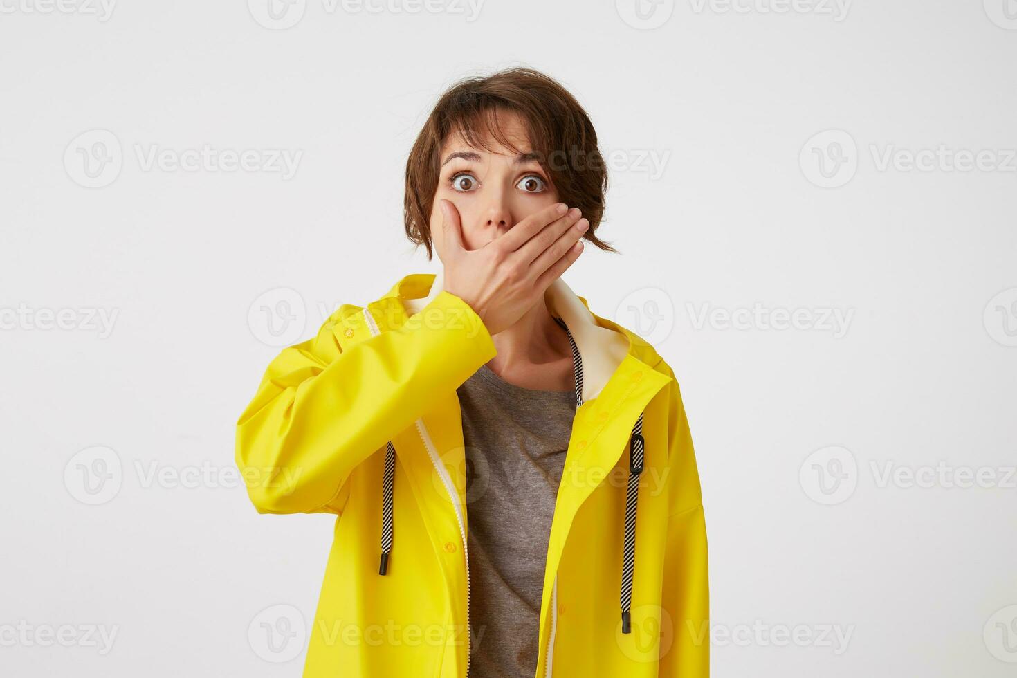 Portrait of short-haired curly woman in yellow rain coat, hears unbelievable news , covered mouth with hand, stands over white wall with wide open eyes with surprised expression. photo