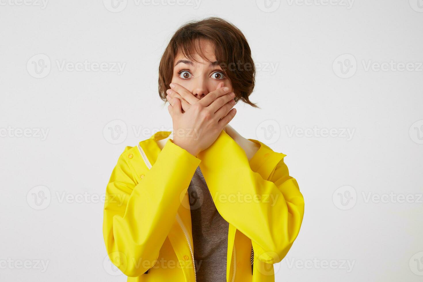 Portrait of scared short-haired curly woman in yellow rain coat, heard a terrible storry, covered mouth with palms, stands over white wall with wide open eyes with afraided expression. photo