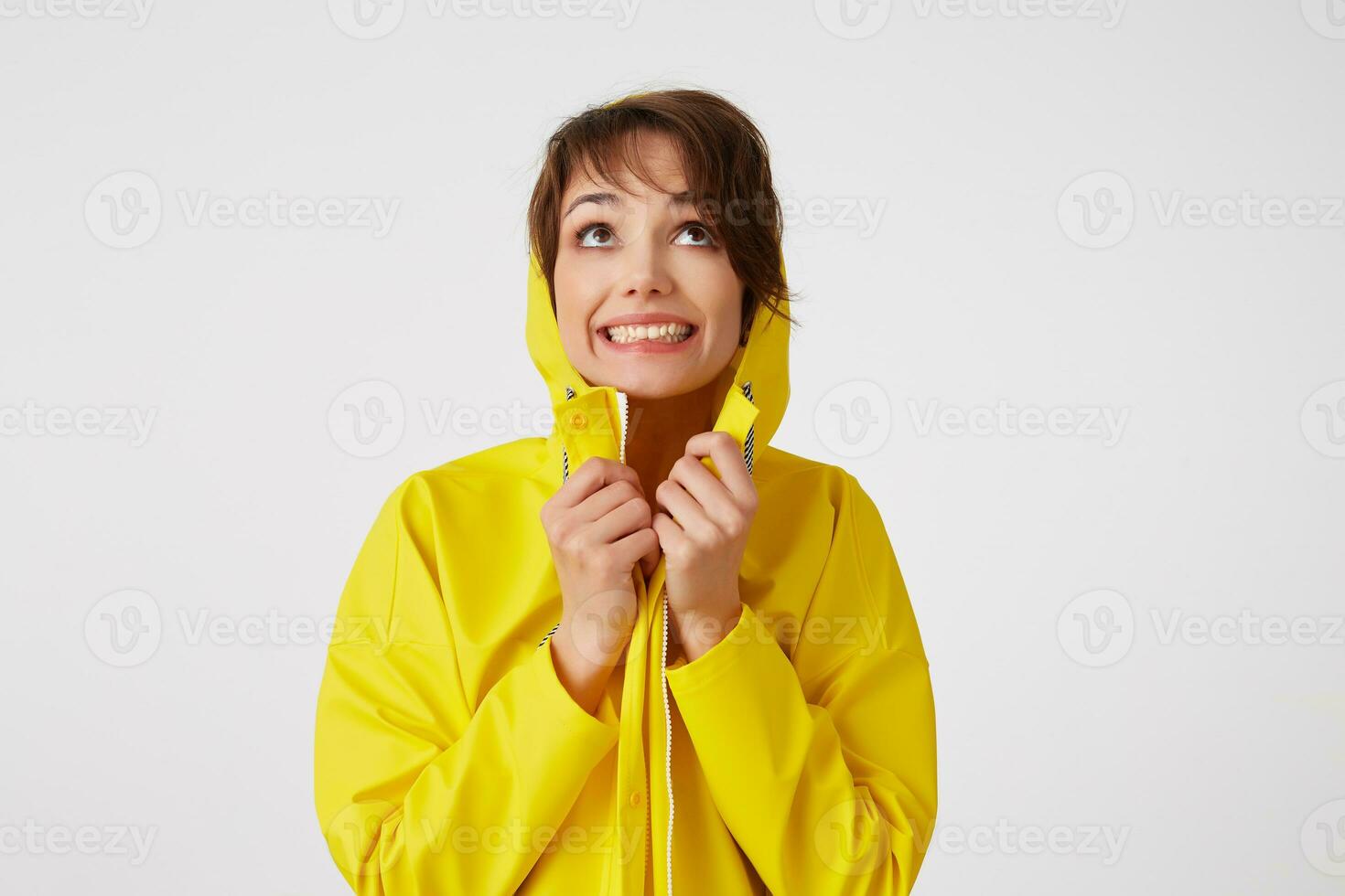 Portrait of young happy cute short haired girl wears in yellow rain coat, hiding under a rain hood, broadly smiles and looks up, stands over white wall. photo