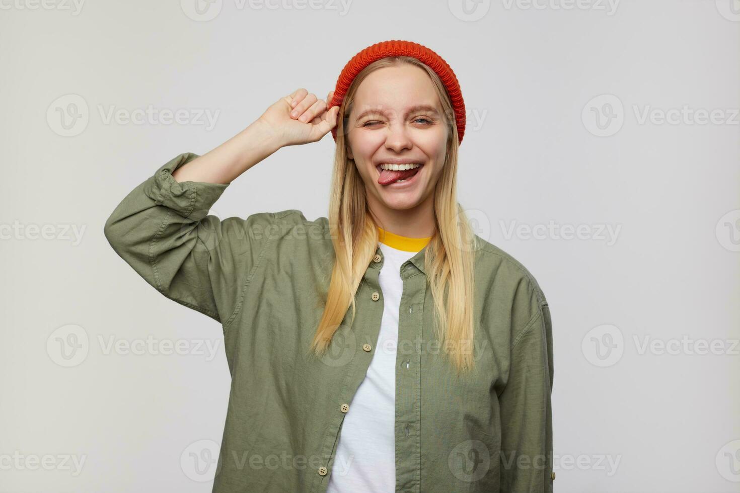 Indoor photo of joyful young attractive white-headed lady with natural makeup winking happily at camera and showing tongue, standing over blue background