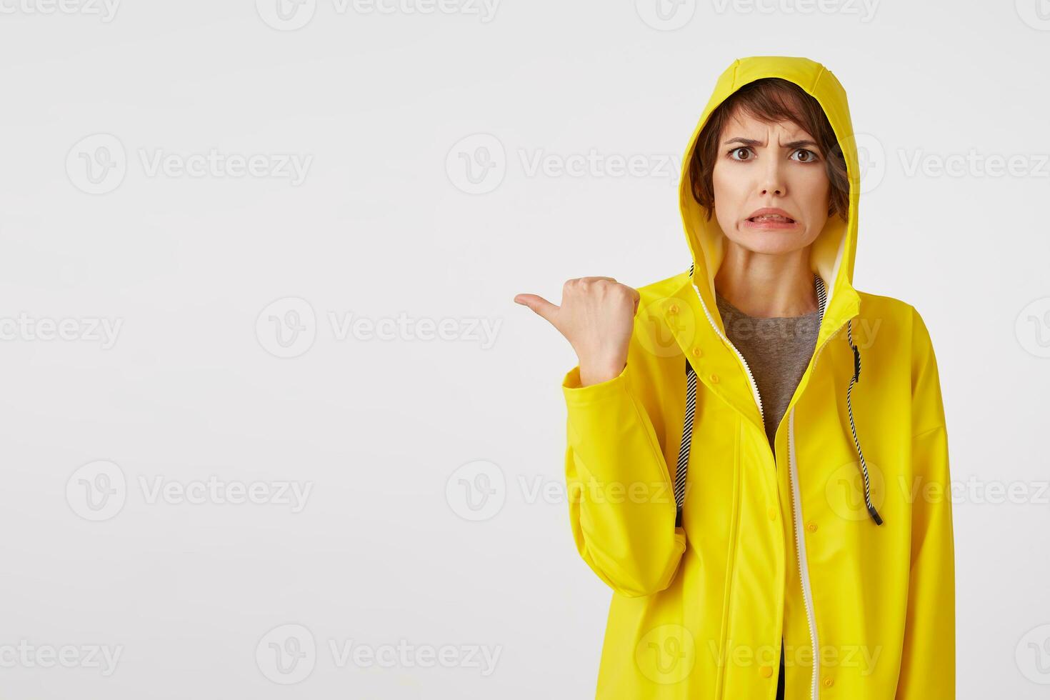 attractive girl in a yellow raincoat with a disgusted expression on her face wants to draw your attention to the copy space on the left pointing with his finger, standing over white wall. photo