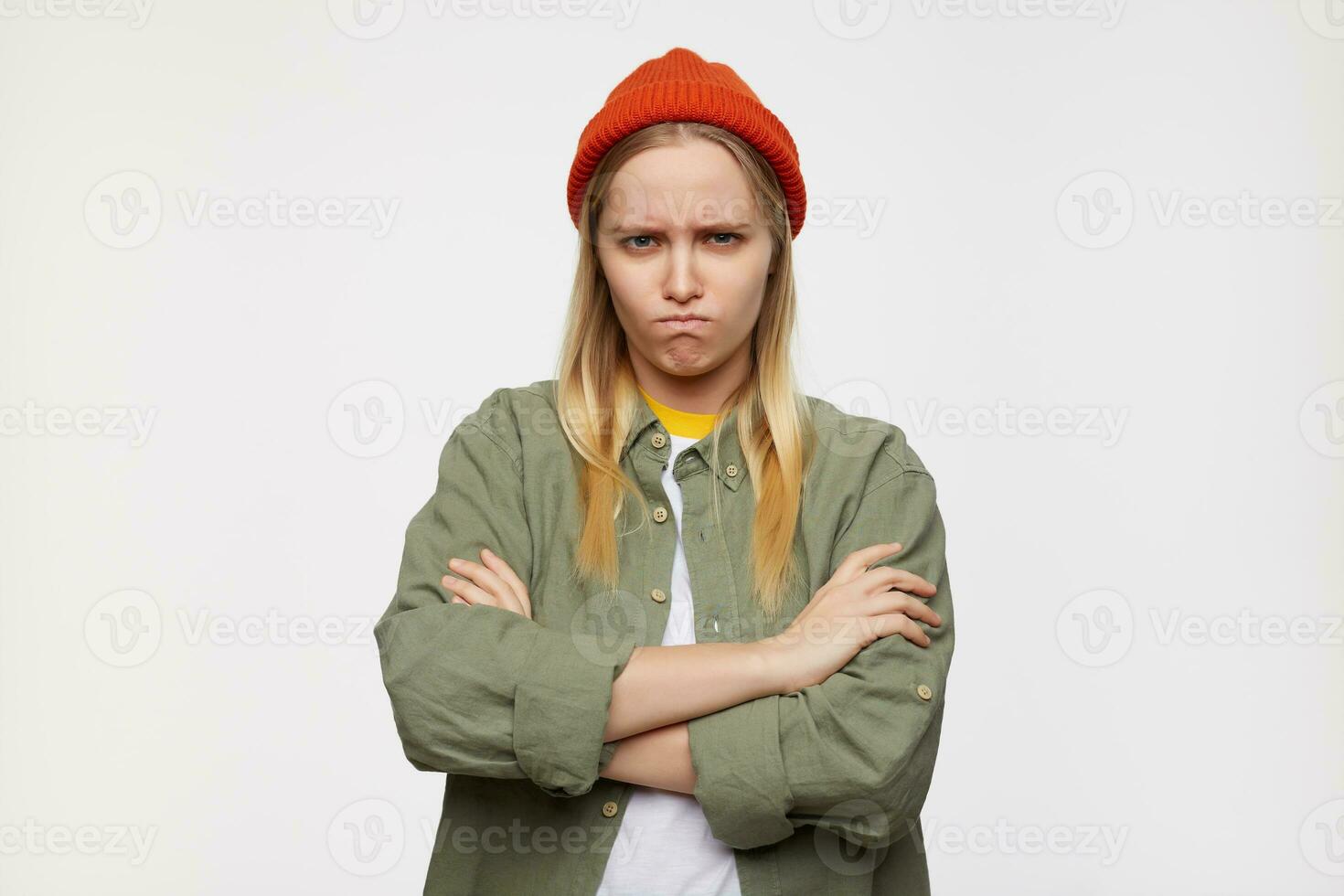 Aggrieved young long haired blonde woman frowning eyebrows while looking severely at camera and folding her hands on chest, posing over blue background photo