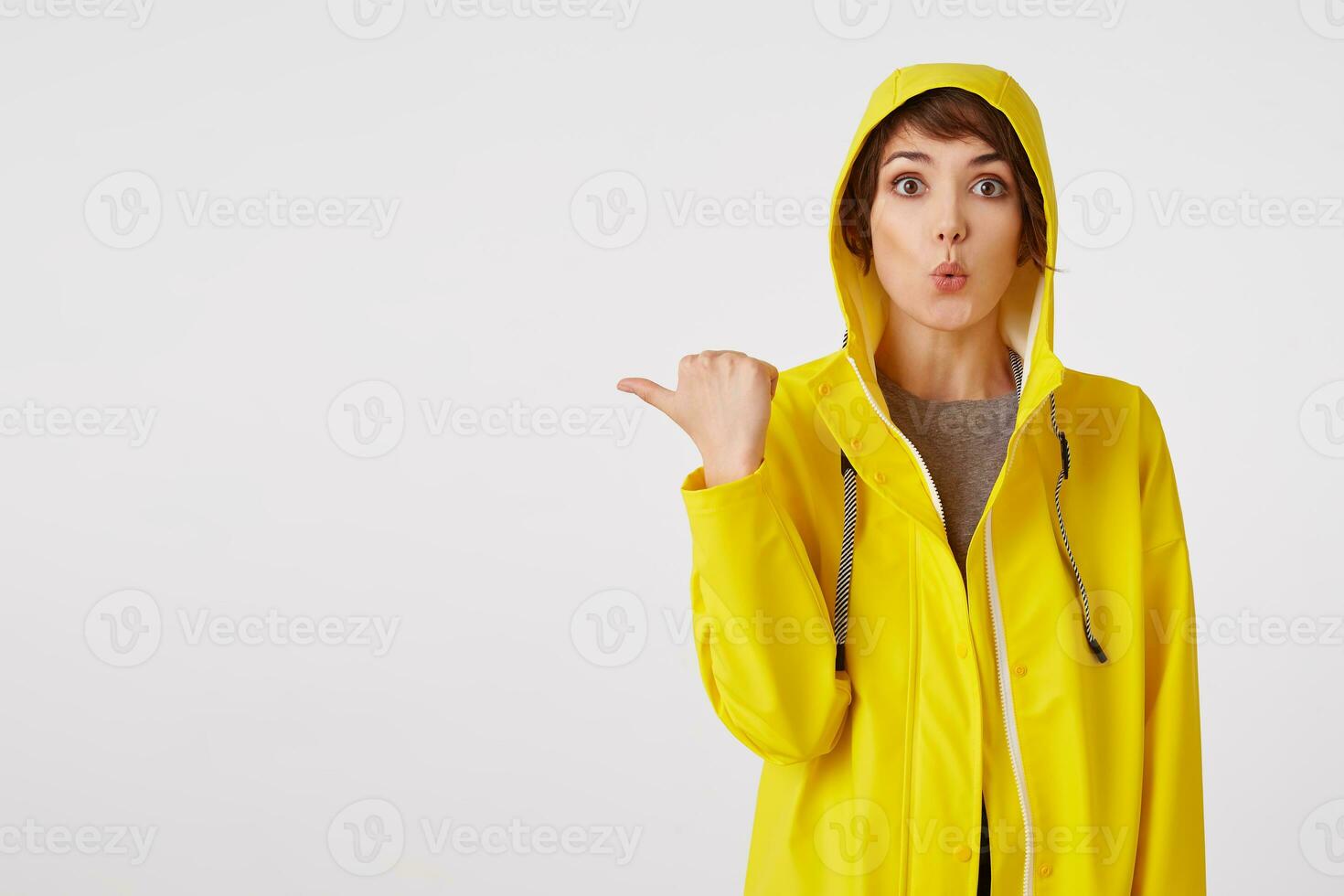 Young attractive girl in a yellow raincoat with a surprised expression on her face wants to draw your attention to the copy space on the left pointing with his finger, standing over white wall. photo