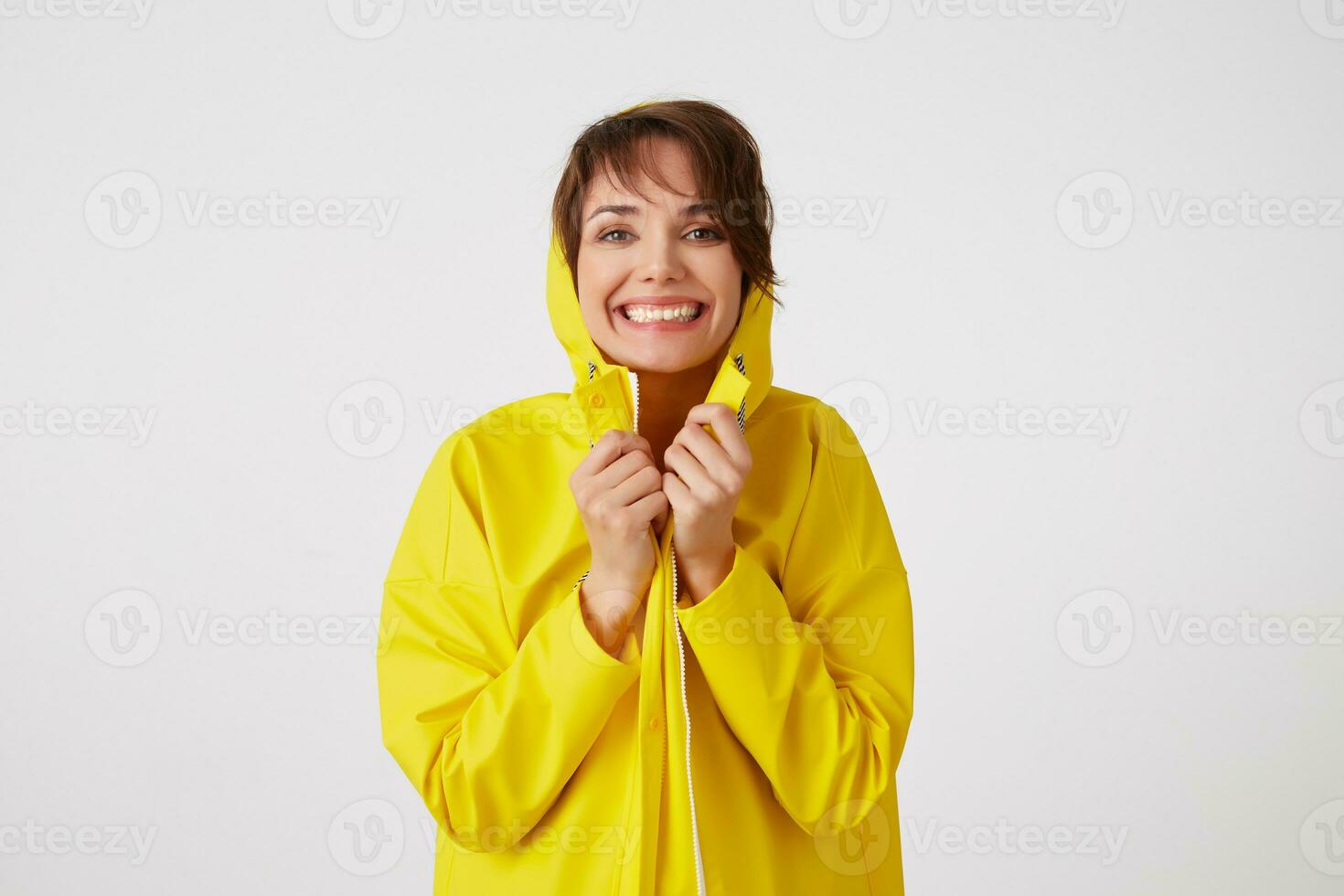 Portrait of young happy cute short haired girl wears in yellow rain coat, hiding under a rain hood, broadly smiles and looks at the camera,stands over white wall. photo