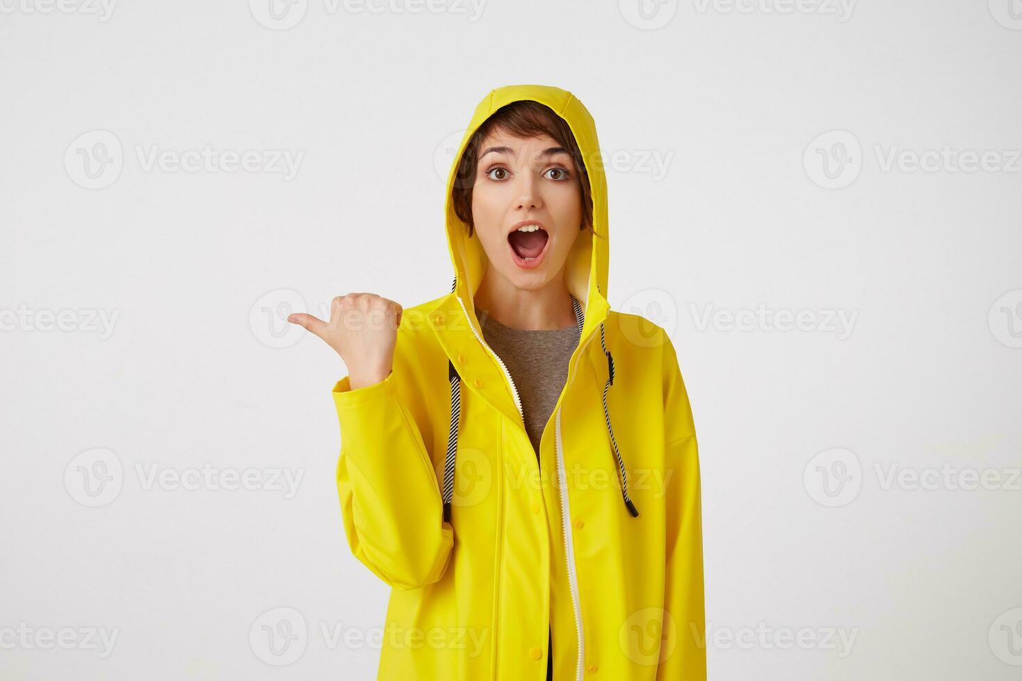 young happy amazed cute short haired girl wears in yellow rain coat, with wide open mouth and eyes, wants to draw you attention and points to copy space at the left, stands over white wall. photo