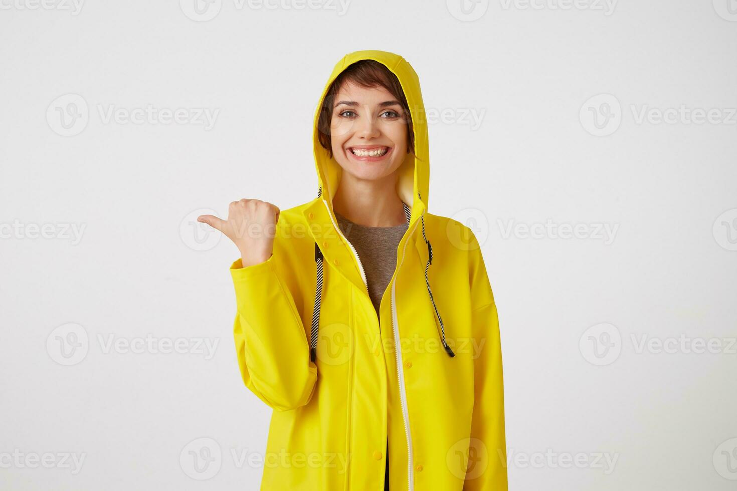 Portrait of young happy cute short haired girl wears in yellow rain coat, broadly smiles, wants to draw you attention and points to copy space at the left, stands over white wall. photo