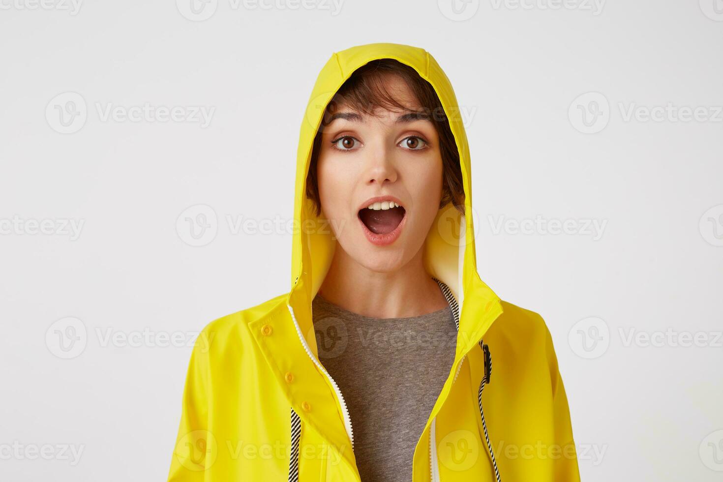 Close up of young attractive girl in a yellow raincoat with a surprised expression on her face, standing over white wall with wide open moth and eyed. Positive emotion concept. photo