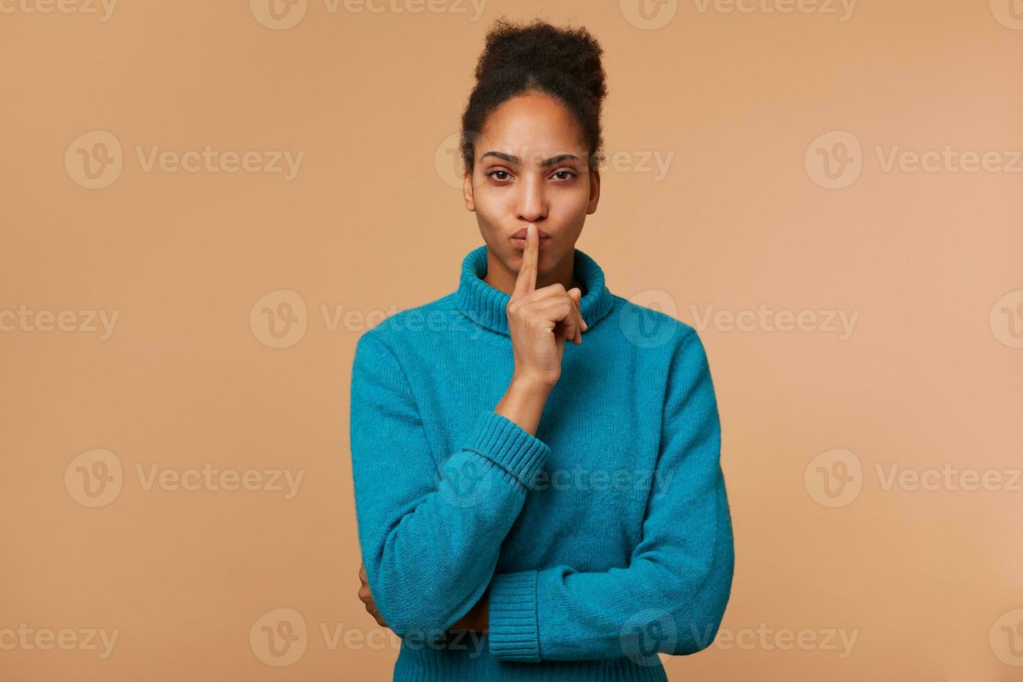 Mysterious African American young lady, demonstrates a gesture of silence, holding an index finger near the mouth calls for privacy to be kept, secret, stay quiet,calm. Isolated over beige background. photo
