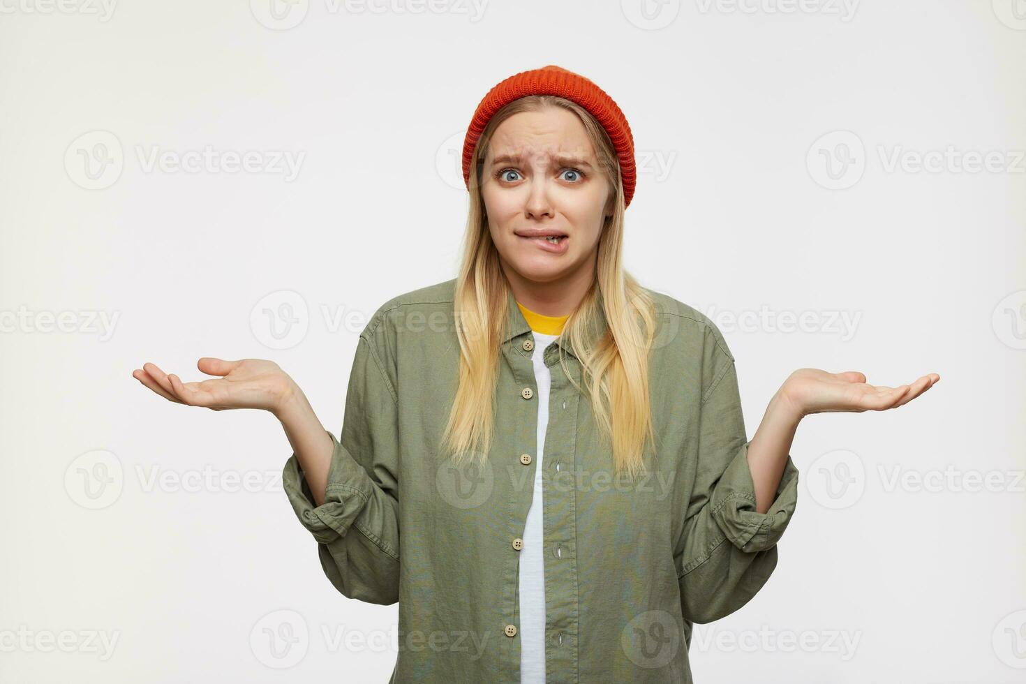 Confused young blue-eyed white-headed female with natural makeup biting worringly underlip and keeping her palms raised, isolated over blue background photo