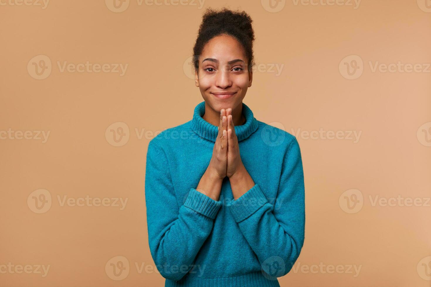 Portrait of young African American girl with curly dark hair wearing a blue sweater. Smiling, keeps palms together, pleads for mercy. Isolated over beige background. photo