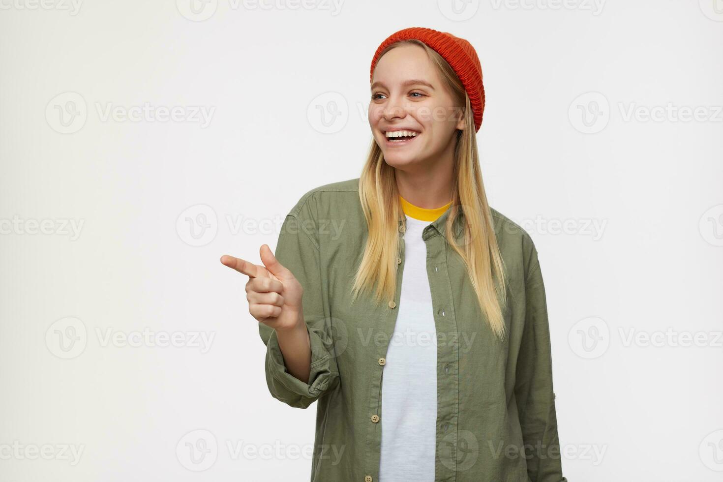 Indoor shot of young lovely long haired blonde woman laughing cheerfully while pointing aside with index finger, standing against blue background in casual clothes photo