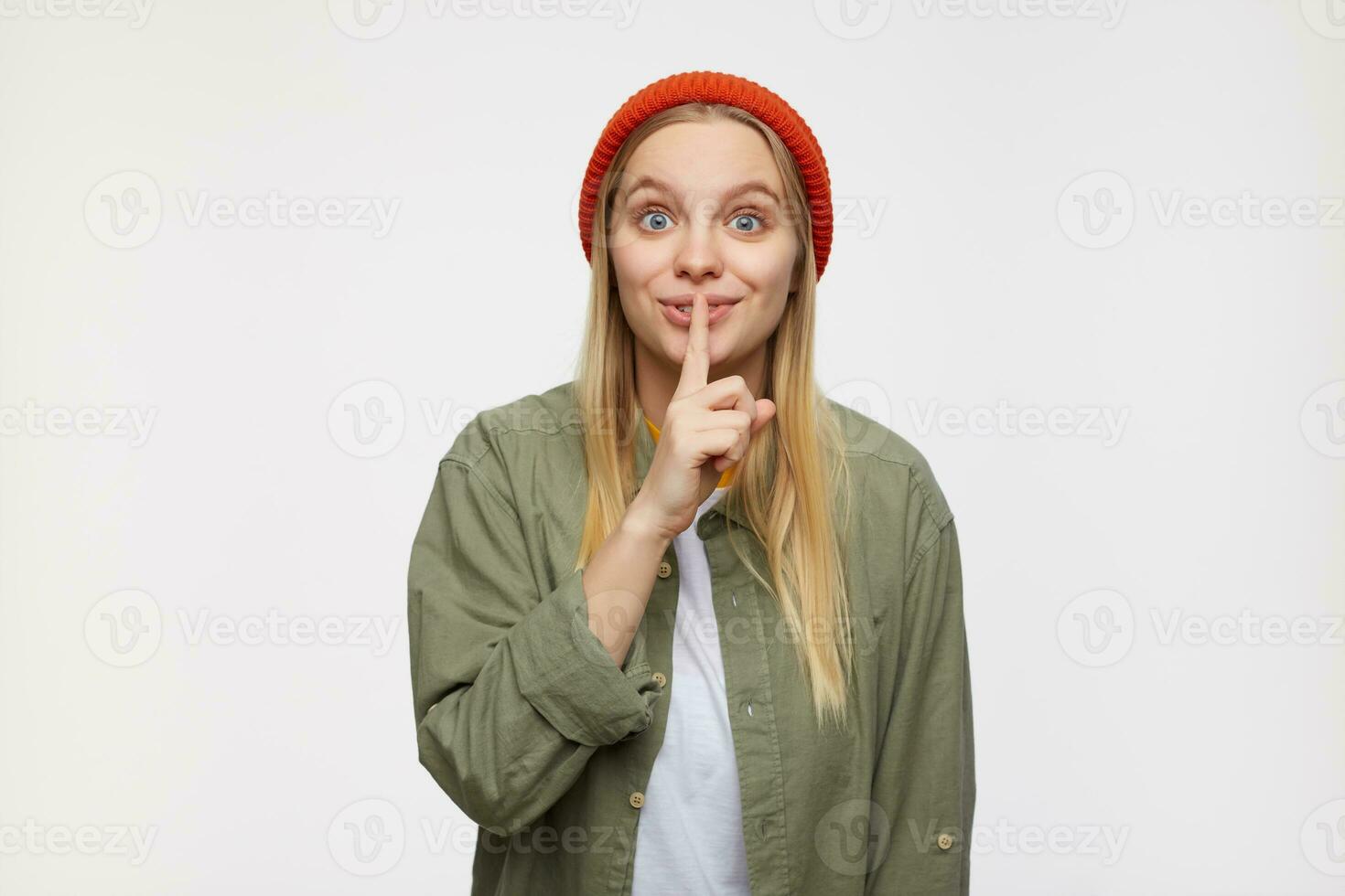 Positive young pretty long haired blonde female with casual hairstyle looking excitedly at camera and raising hand with hush gesture, isolated over blue background photo