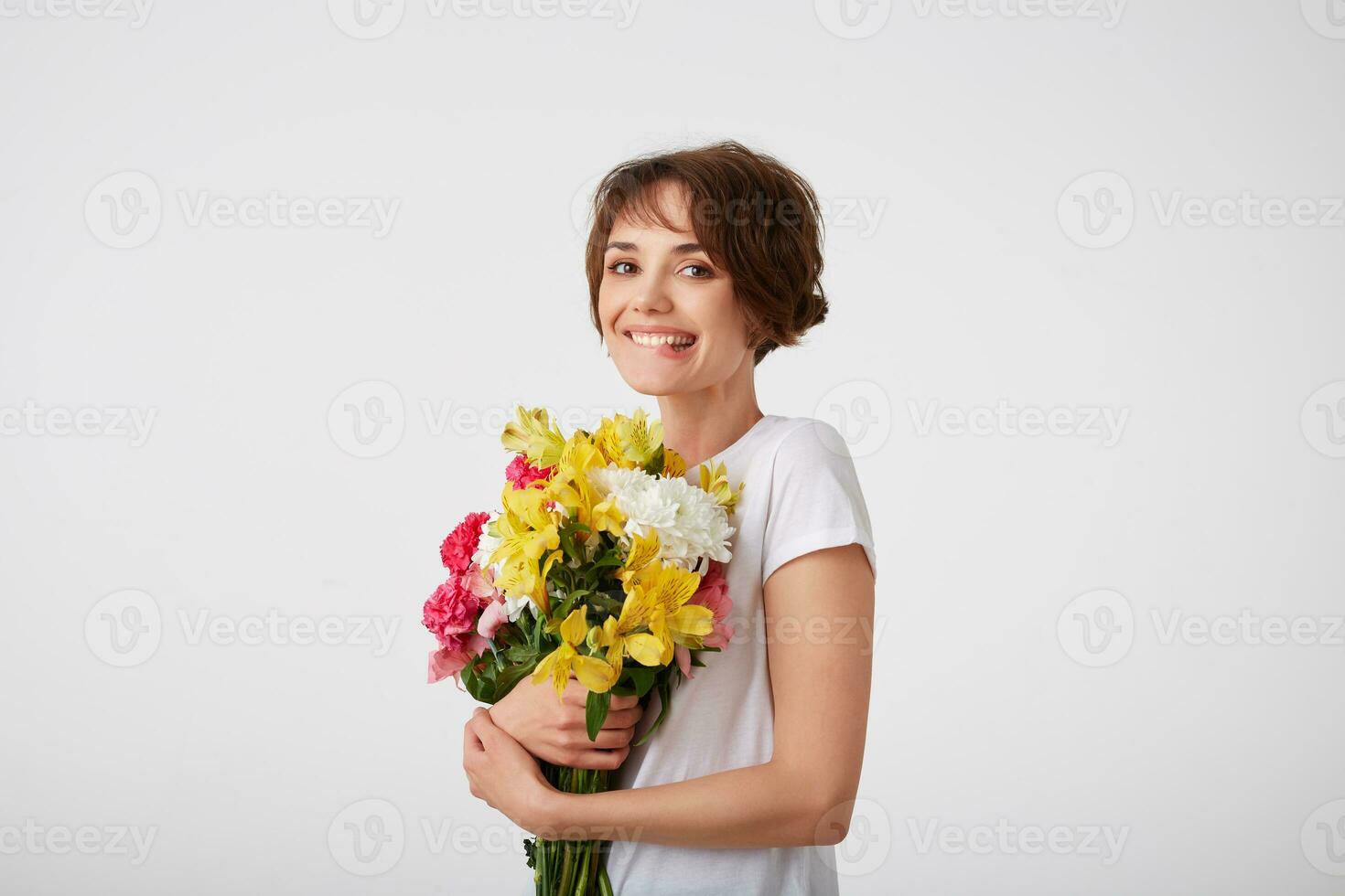 Young smiling cute short haired girl in white blank t-shirt, holding a bouquet of colorful flowers, very glad to such a gift from her boyfriend, bites his lip and smiling over white wall. photo