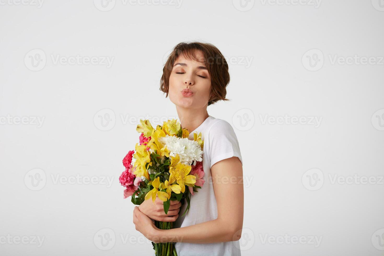 Portrait of young lovely cute short haired lady in white t-shirt, holding a bouquet of colorful flowers, very glad to such a gift from her boyfriend, sends a kiss with closed eyes over white wall. photo