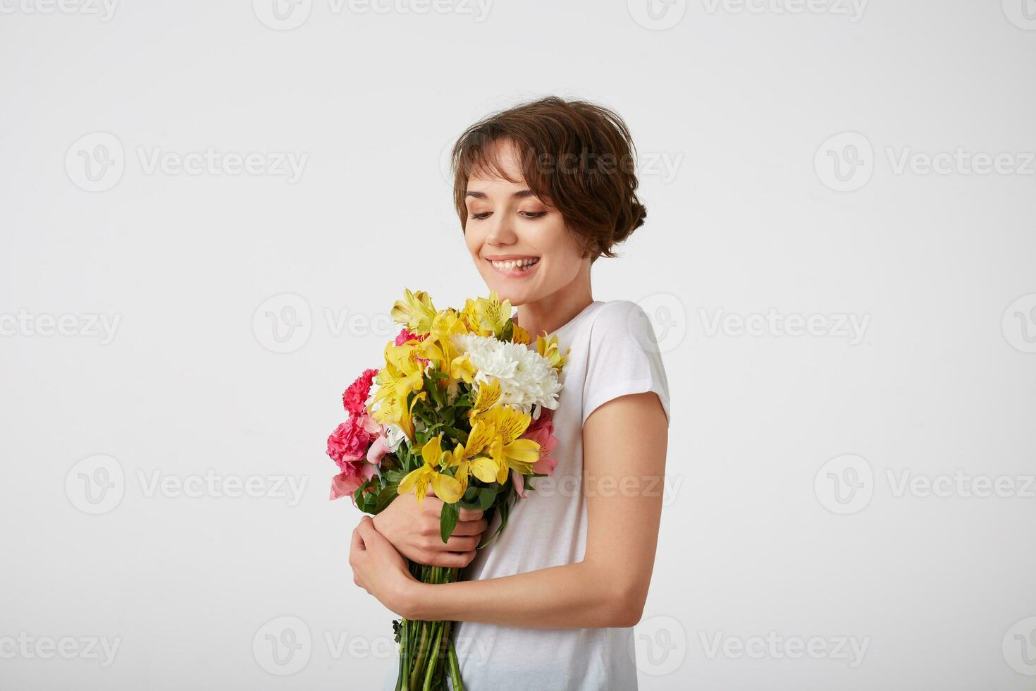 Cute happy young short haired girl in white blank t-shirt, holding a bouquet, bites his lip and looks at the of colorful flowers, enjoying the smell, standing over white background. photo