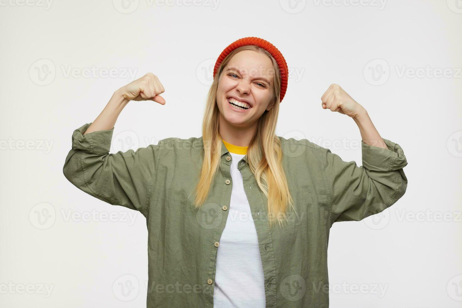 Portrait of young joyful long haired blonde lady frowning cheerfully her face and keeping her hands raised, posing over blue background in casual clothes and red hat photo