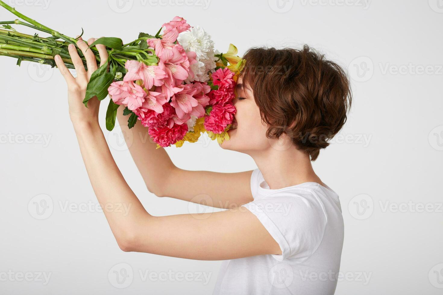 Portrait of nice short haired girl in white blank t-shirt, holding a bouquet of colorful flowers, enjoying the smell, standing over white background with closed eyes. photo