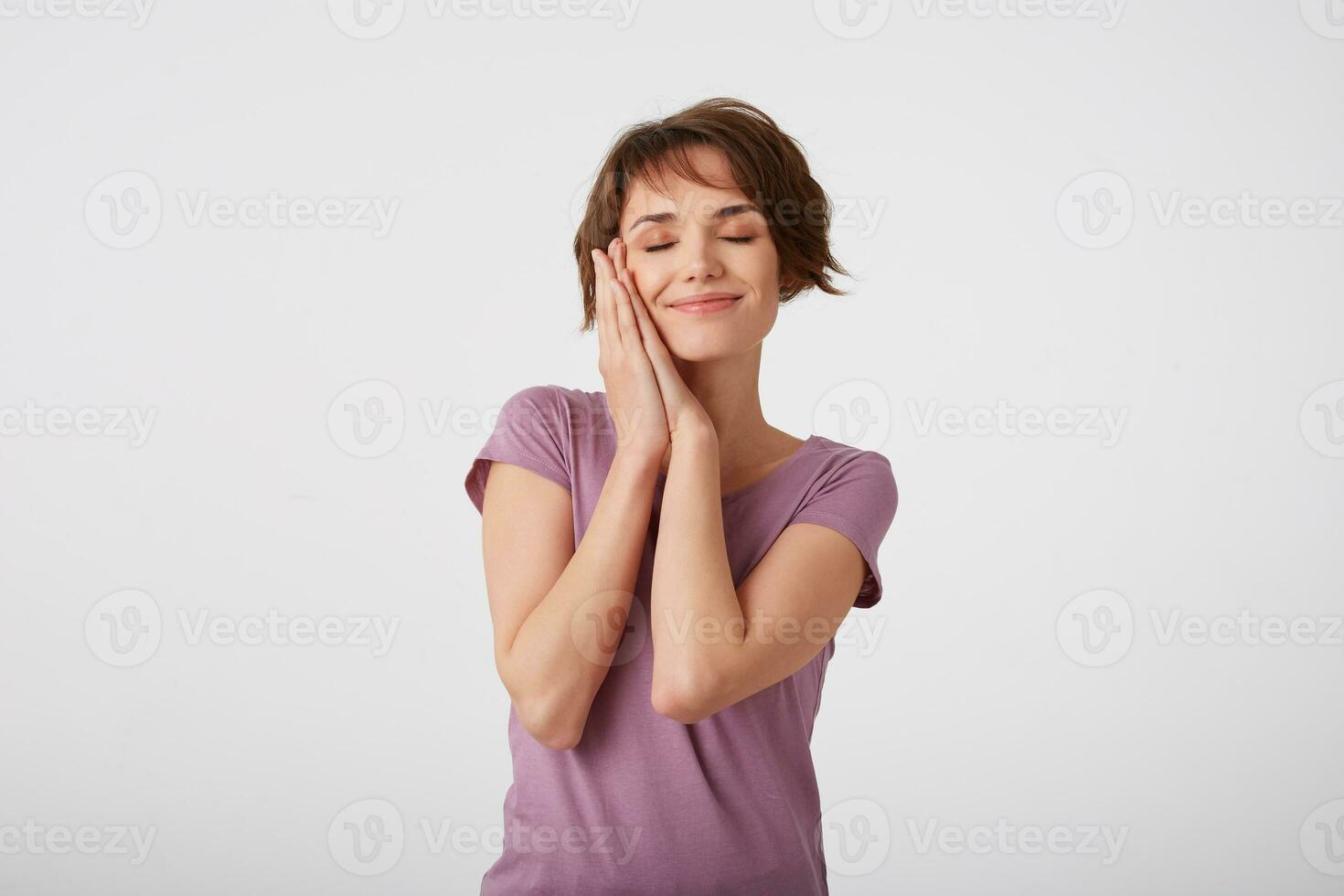 Young attractive young short-haired lady in blank t-shirt, pretends sleeping, leans on hands, has tender smile, takes nap. Standing over white background. photo