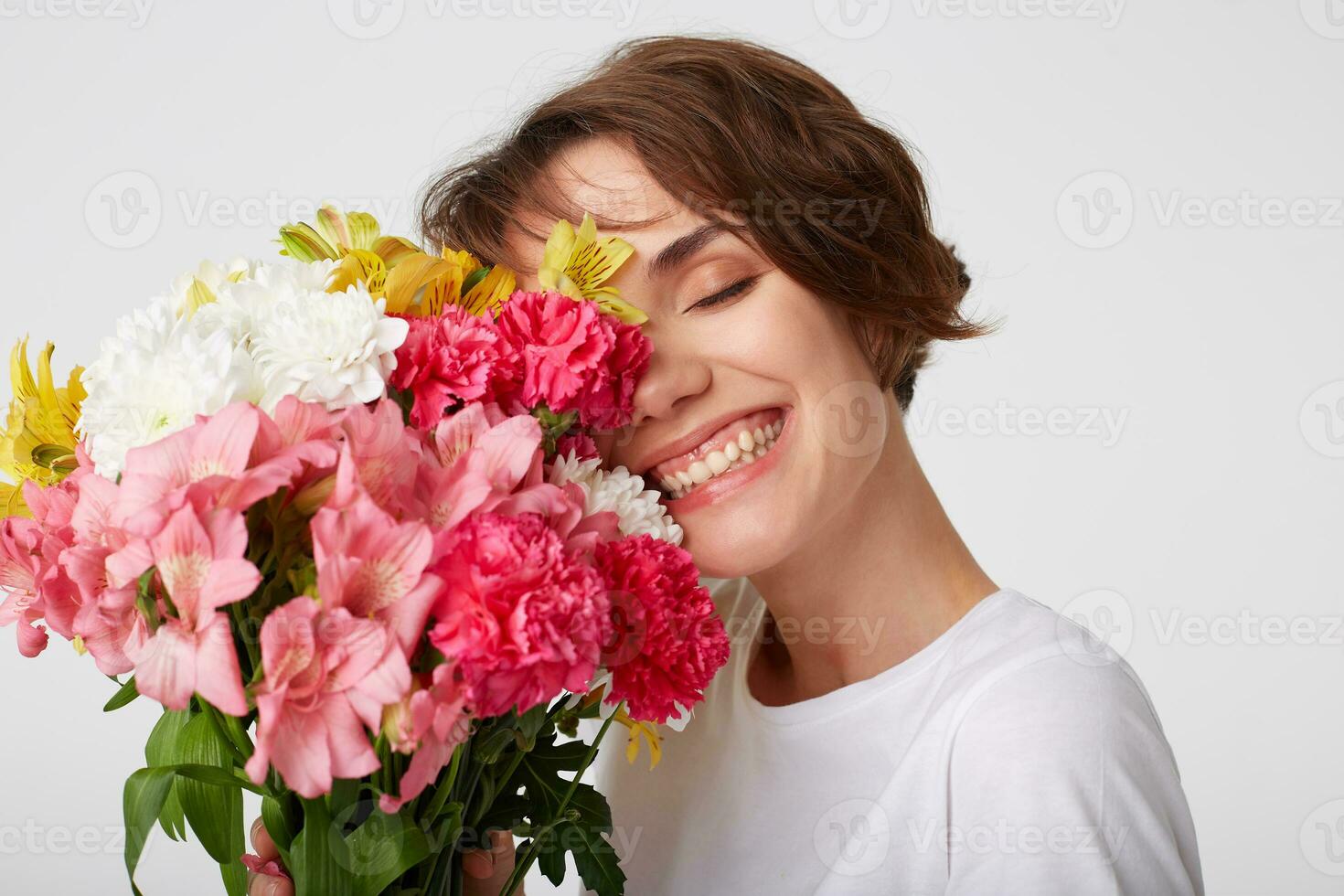 Portrait of nice short haired girl in white blank t-shirt, holding a bouquet, covers face with flowers, standing over white background with closed eyes. photo