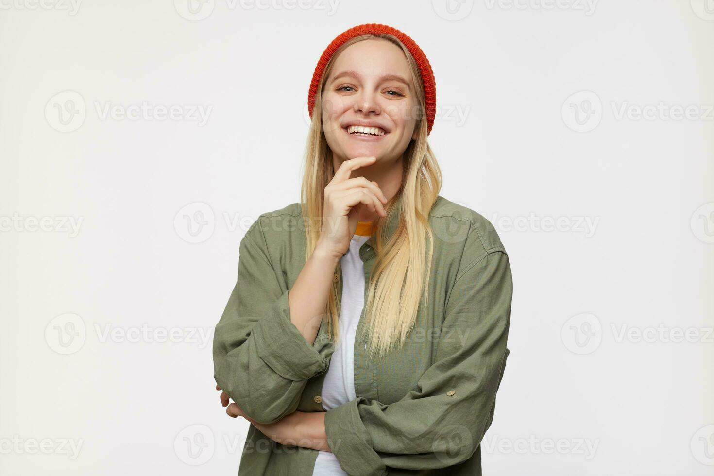 Studio photo of charming young white-headed female looking cheerfully at camera with wide smile and touching her chin with raised hand, isolated over blue background
