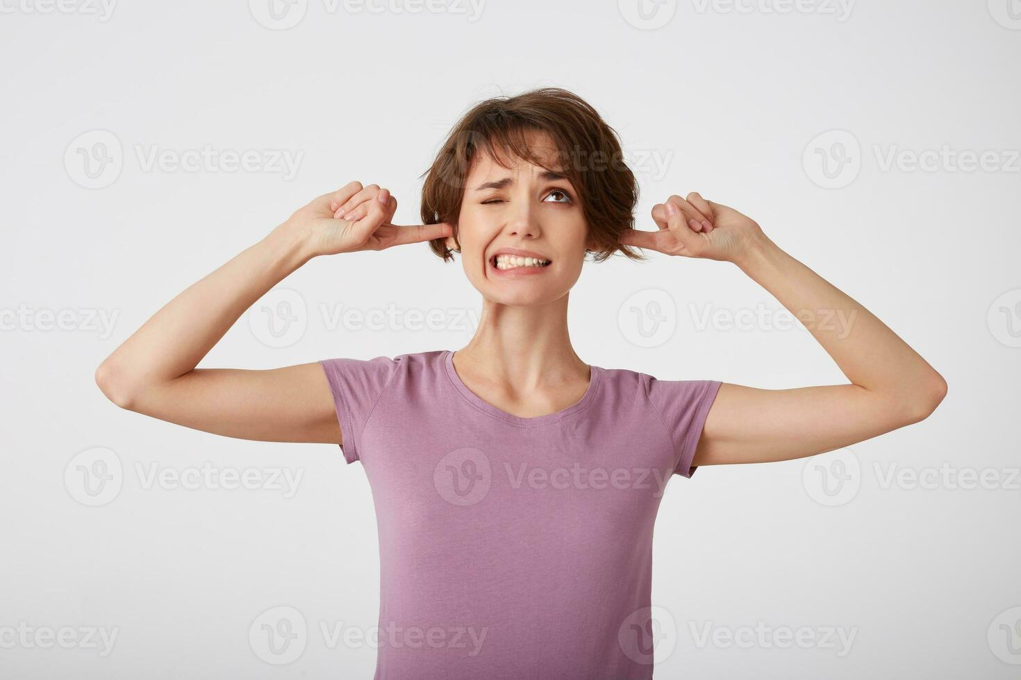 Young nice doubted short-haired lady in blank t-shirt, unwills hear bothering noise, plugs earholes with index fingers, clenches teeth and frowns face in dissatisfaction. Stands over white wall. photo