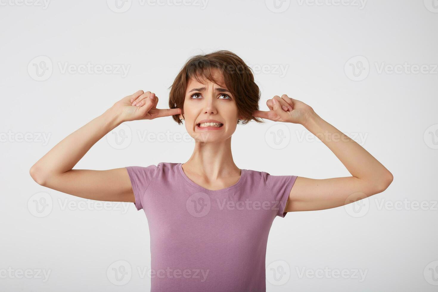 Young nice doubted short-haired lady in blank t-shirt, plugs ears as hears awful noise or music, squints face, avoids loud sound, ignores something unpleasant. Stands over white wall. photo