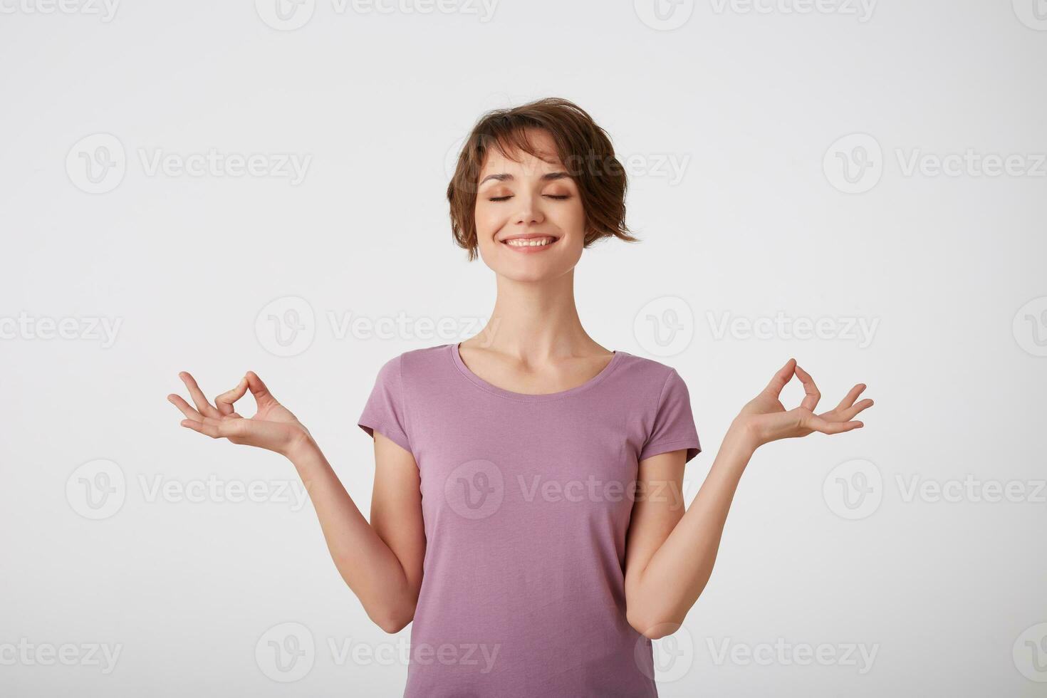 Concentrated young attractive young short-haired lady in blank t-shirt, keeps hands in okay gesture, meditates indoor, has eyes closed, tries to relax, isolated on white wall. photo