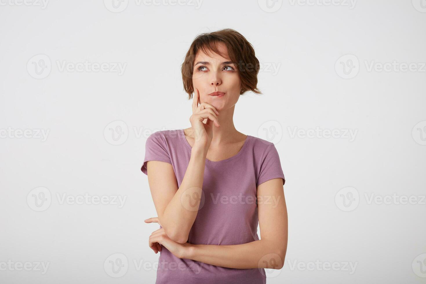 Young attractive young short-haired woman in blank t-shirt looks aside, thinks about rest on weekened, what to buy. Stands over white background. photo