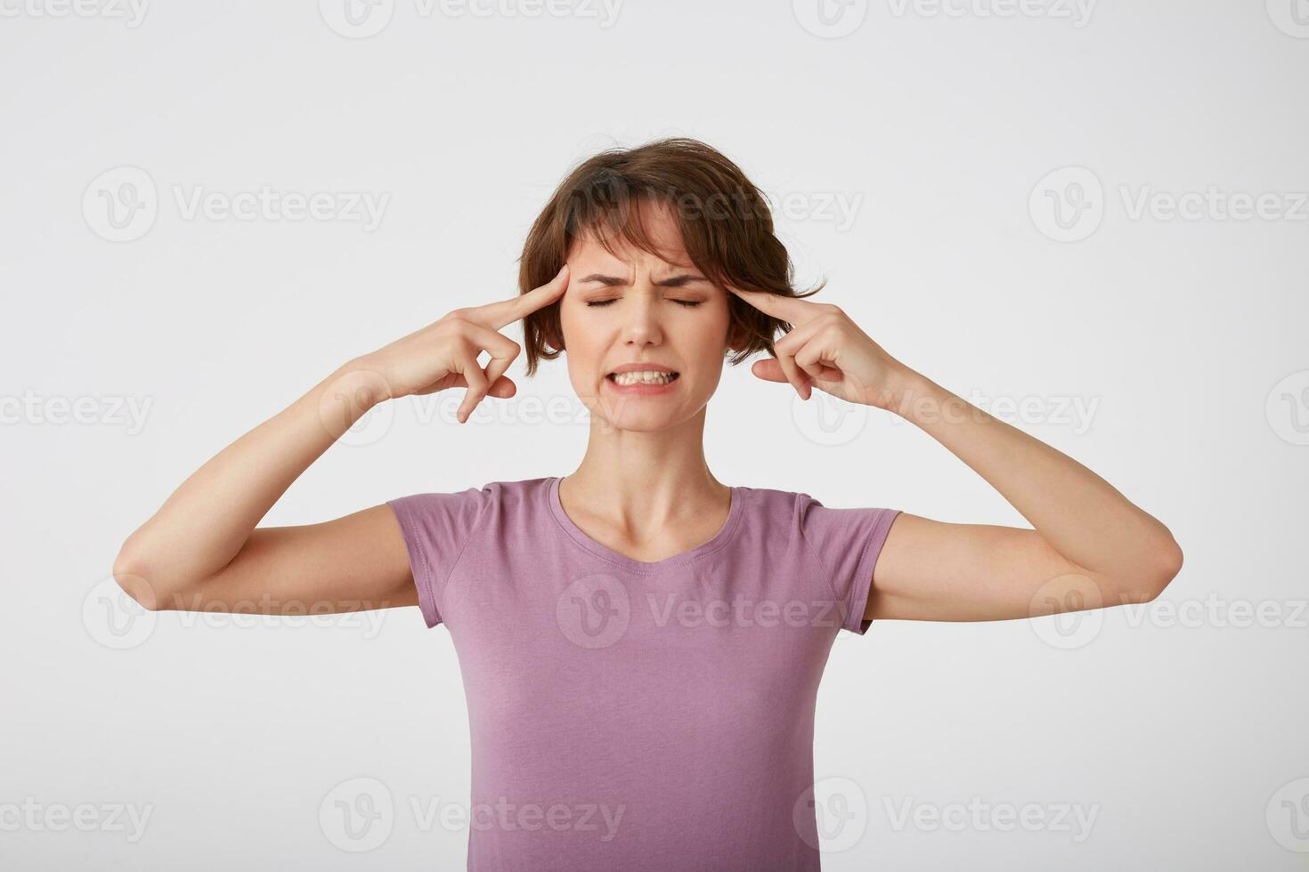 Young nice short-haired lady in blank t-shirt, suffers from migraine, cannot hold pain, touches temples, needs painkillers, clenches teeth, feels discomfort isolated over white wall. photo