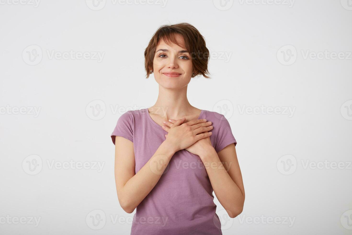 Beautiful cheerful short-haired lady in blank t-shirt, heard a nice compliment, smiling with crossed palms on heart, stands over white background. Positive Emotion concept. photo