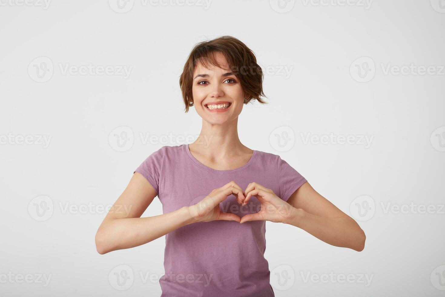 hermosa alegre de pelo corto dama en blanco camiseta, muestra corazón forma gesto, usa casual t camisa, siendo contento, soportes terminado blanco antecedentes. amor símbolo, romántico humor. foto