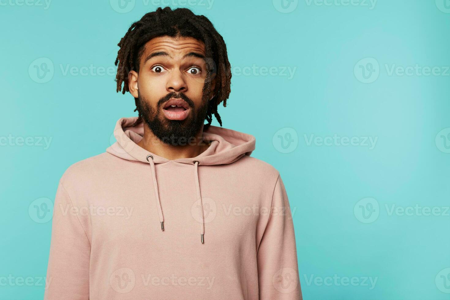 Bemused young pretty brunette bearded man with dark skin looking amazedly at camera with wide eyes and mouth opened while posing against blue background photo