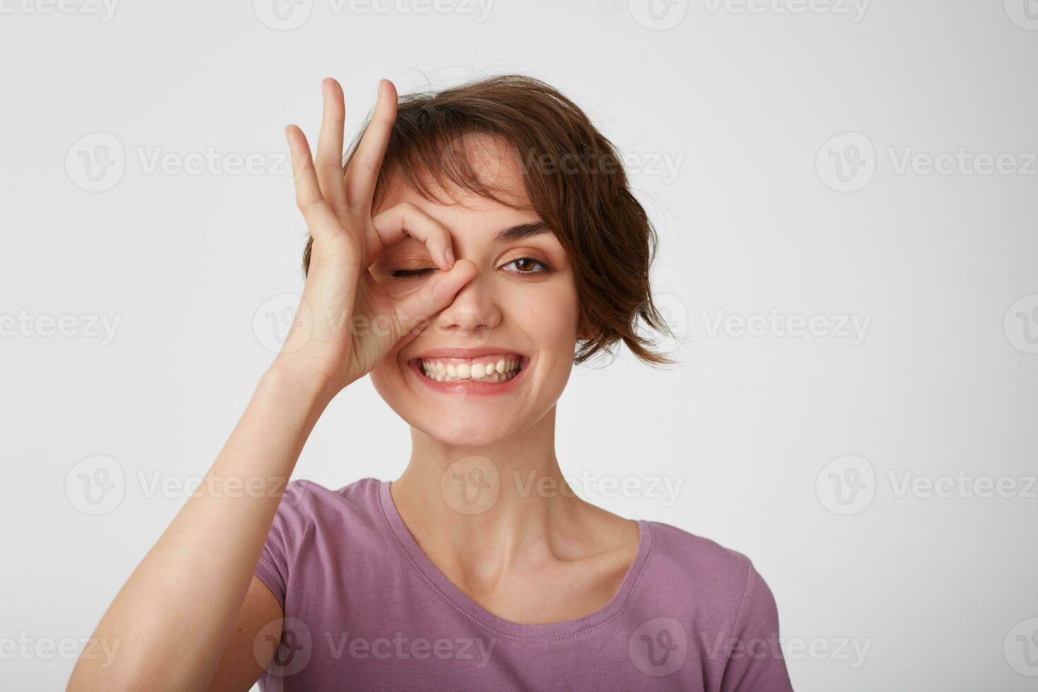 Close up of young attractive happy short-haired lady in blank t-shirt, standing over white wall and looking through okay gesture and broadly smiling Positive emotion concept. photo