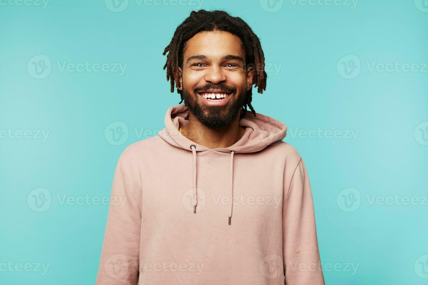 Cheerful young lovely brunette bearded man with dark skin smiling widely while looking happily at camera, keeping hands down while posing over blue background photo