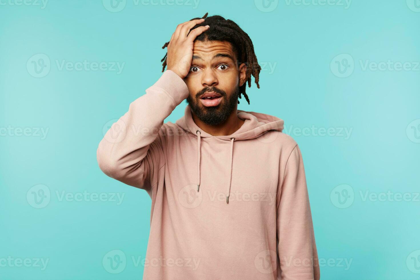 Studio photo of puzzled young handsome brunette bearded male holding raised palm on his head while looking confusedly at camera, isolated over blue background