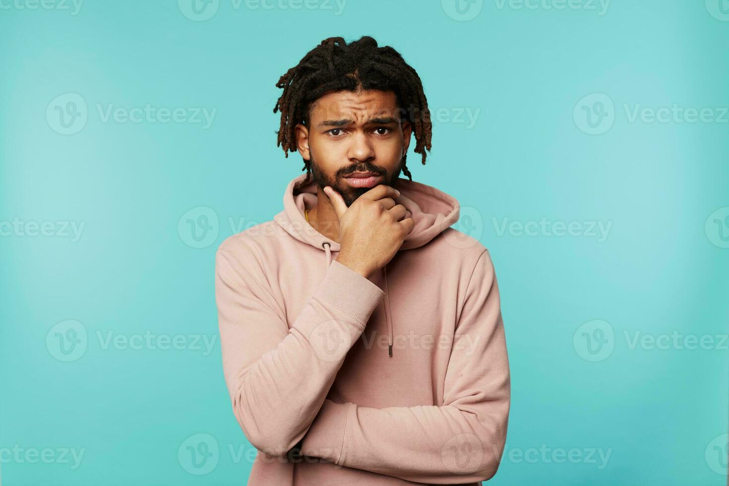 Puzzled young dark haired bearded guy with dreadlocks holding his chin with raised hand while looking pensively at camera, standing over blue background photo